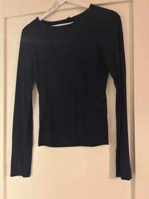 Mörkblå långärmad topp - En enkel och stilren mörkblå långärmad topp med rund halsringnin och slit vid hermens slut är i st S. Toppen har en slim passform och är tillverkad i ett mjukt och stretchigt material som sitter snyggt på kroppen. Perfekt att matcha med jeans eller kjol.