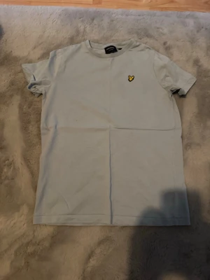Ljusgrå Lyle & Scott t-shirt - Snygg ljusgrå t-shirt från Lyle & Scott med klassisk gul logga på bröstet. T-shirten har rund halsringning och korta ärmar. Perfekt för en stilren och enkel look. Materialet är mjukt och bekvämt bomullstyg.
