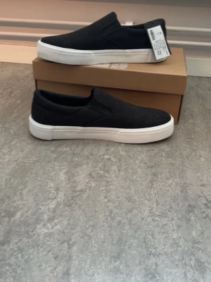 Svarta slip-on sneakers från 157 - Svarta slip-on sneakers från 157 med vit sula och enkel, stilren design. Ovandelen är i textil och skon har elastiska paneler på sidorna för enkel påtagning. Perfekta för en clean och avslappnad look.