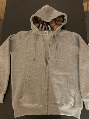 Grå zip hoodie med Burberry-mönster - Snygg grå zip hoodie från Burberry med klassiskt rutigt mönster i huvan. Hoodien har dragsko i huvan, två stora fickor fram och ribbade muddar. Tillverkad i mjuk bomullsblandning som känns skön mot huden. Perfekt för en avslappnad och trendig look.