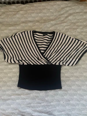 Randig omlottopp i svart och vitt - Snygg omlottopp med breda, korta ärmar och v-ringning. Toppen har svart och vitt randigt mönster upptill och enfärgad svart midja. Ribbad och croppad passform, perfekt till jeans eller kjol.