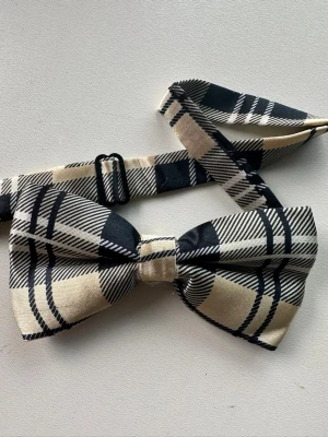 Beige och svart rutigt fluga - Stilig fluga med justerbart band i rutigt mönster i beige och svart. Flugan har ett förknutet utförande och stängs med hake. Passar som accessoar till skjorta och kostym eller andra mer uppklädda tillfällen. Materialet är slätt, vävt sarin med lätt glansig yta. Oanvänd, ny i förpackning. 