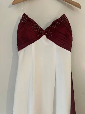 Vit och vinröd långklänning med pärlor - Elegant långklänning i vit och vinröd färg med pärldetaljer på överdelen. Klänningen har smala axelband och ett vackert fall. Perfekt för dig som vill sticka ut med en stilren och lyxig look. Passar till bal och bröllop