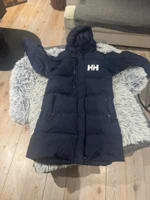 Mörkblå lång dunjacka från Helly Hansen - Lång mörkblå dunjacka från Helly Hansen med huva och vit HH-logga på bröstet. Jackan har dragkedja framtill och två sidofickor med dragkedja. Perfekt för kalla dagar och har en quiltad design som håller dig varm.