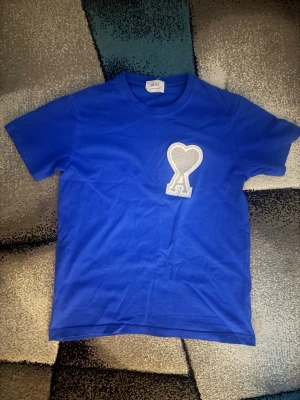 Blå t-shirt från Ami med hjärtdetalj - Snygg blå t-shirt från Ami med ett stort vitt hjärta broderat på bröstet. Klassisk passform och rund halsringning. Perfekt för dig som vill ha en enkel men ändå unik look. Tröjan är i storlek M men passar också bra i S