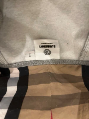 Grå Burberry hoodie med klassiskt foder - Säljer en grå hoodie från Burberry med det ikoniska rutiga fodret i beige, svart, vitt och rött. Hoodien har huva och dragkedja framtill. Perfekt för dig som vill ha en stilren och lyxig look med klassiska Burberry-detaljer. Storlek s-m