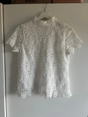 Vit spetsblus med kort ärm - Säljer en superfin vit blus i spets med korta ärmar och hög hals. Blusen har ett detaljerat spetsmönster och knapp i nacken. Perfekt för dig som gillar romantisk och elegant stil.