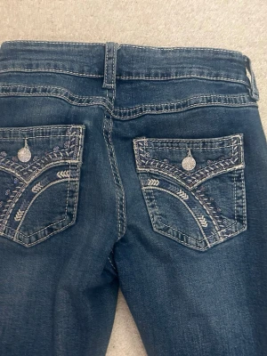 Blå bootcut jeans med broderade fickor - Snygga blå bootcut jeans med broderade bakfickor och dekorativa silverknappar. Jeansen har klassisk femficksdesign och coola detaljer med ljusa broderier på fickorna. Perfekt för dig som gillar en unik och trendig look.