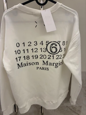 Maison Margiela tröja  - Vit hoodie från Maison Margiela Paris med det ikoniska siffertrycket och logga på bröstet. Oversized passform och ribbade muddar. Perfekt för dig som gillar streetstyle och exklusiva märken.