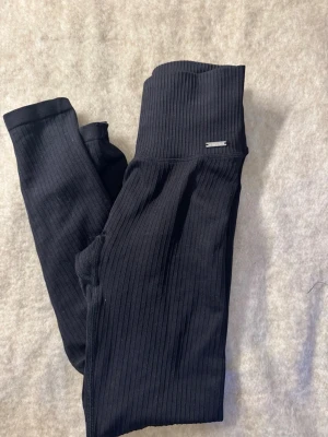 Svarta ribbade leggings Aim'n - Säljer ett par svarta ribbade leggings från Aim'n med hög midja och stretchigt material. De har en diskret logga framtill och sitter tight för en snygg siluett. Perfekta för både träning och chill.