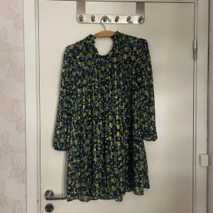 Blommig miniklänning med volangkrage - Kort klänning med blommigt mönster i blått, gult och grönt. Har lång ärm, volangkrage och lätt rynkad överdel. Materialet ser ut att vara ett tunt, mjukt tyg som faller fint. Perfekt för dig som gillar färgglada och lekfulla plagg.