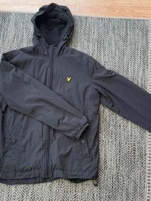 Svart jacka Lyle & Scott - Snygg svart vindjacka från Lyle & Scott med huva och dragkedja framtill. Jackan har två fickor med knappstängning och det klassiska gula logomärket på bröstet. Tillverkad i lätt och slitstarkt material med meshfoder för extra komfort.
