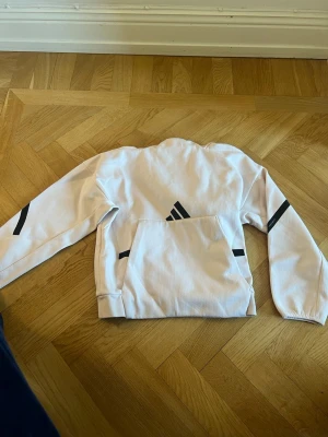 Vit hoodie från Adidas med svarta detaljer - Säljer en vit hoodie från Adidas med svarta detaljer på ärmarna och logga på bröstet. Hoodien har en stor magficka och ribbade muddar vid ärmslut och nederkant. Tillverkad i mjukt material som känns skönt mot huden. Perfekt för en sportig och avslappnad stil.