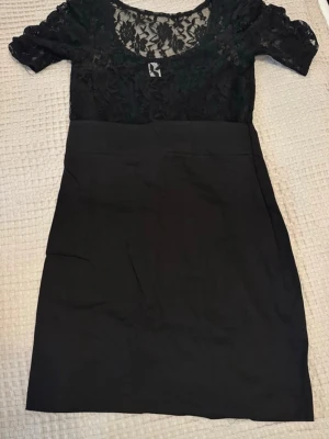 Svart klänning med spetsdetaljer - Snygg svart klänning med korta ärmar och överdel i transparent spets. Rund halsringning och figurnära passform upptill, med slät kjoldel. En fin slits där bak. I storlek M. Aldrig använts. 🌸
