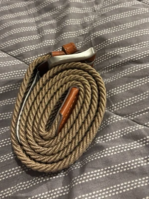 Beige flätat bälte med metallspänne - Snyggt beige flätat bälte med detaljer i brunt skinn och ett stilrent metallspänne. Bältet har en bred och mjuk design som gör det både bekvämt och trendigt. Perfekt accessoar för att lyfta din outfit.