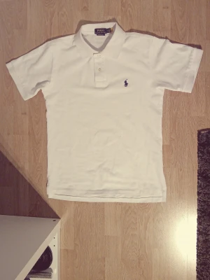 Vit pikétröja från Ralph Lauren - Klassisk vit pikétröja från Ralph Lauren med broderad logga på bröstet. Tröjan har krage, korta ärmar och knäppning framtill. Tillverkad i bomull för en skön och luftig känsla. Storlek S men passar även XS.