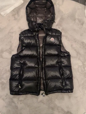 Svart dunväst från Moncler med huva - Snygg svart dunväst från Moncler med avtagbar huva och två dragkedjefickor. Västen har glansig finish och klassisk Moncler-logga på bröstet. Perfekt för lager-på-lager och streetstyle. Materialet är dunfyllt och yttermaterialet är glansigt syntet.