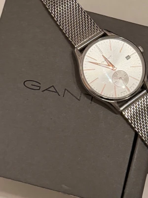 Silverfärgad klocka från GANT - Stilren klocka från GANT med mesh-armband i silverfärgat stål. Urtavlan är ljus med minimalistiska detaljer och datumvisning. Klockan har en rund form och klassisk design som passar till många olika stilar. Köpt för 1890
