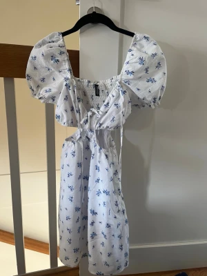 Vit klänning  - Superfin vit klänning med blått blommönster. Klänningen har snörning framtill, cut-out detaljer i midjan och en söt, romantisk stil. Perfekt för varma dagar och somriga vibes.