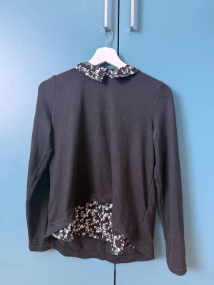 Tröja - Svart långärmad tröja med blommig krage och blommigt tyg i nederkanten baktill. Tröjan har en snygg detalj med den kontrasterande vita blomman mot svart bakgrund. Perfekt för dig som gillar en söt och stilren look.