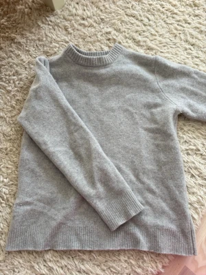 Massimo Dutti tröja - Säljer en stilren ljusgrå stickad tröja från Massimo Dutti. Tröjan har rund halsringning, ribbade muddar och är tillverkad i en mjuk mix av ull och kashmir. Perfekt för dig som gillar klassisk och enkel stil.