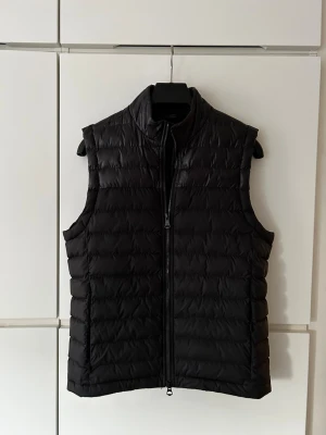 Svart dunväst från J.Lindeberg - Säljer en svart dunväst från J.Lindeberg med quiltad design och hög krage. Västen har dragkedja framtill och är ärmlös, perfekt för lager-på-lager. Materialet är syntet med dunfyllning och logotyp på bröstet.