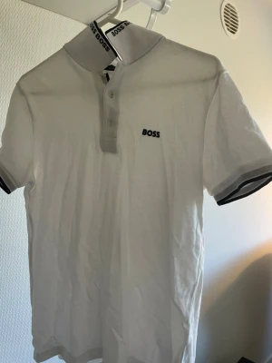 Vit pikétröja från BOSS - Klassisk vit pikétröja från BOSS med korta ärmar och svart logga broderad på bröstet. Tröjan har krage med BOSS-tryck på insidan och knappar framtill. Perfekt till jeans eller shorts för en clean look.