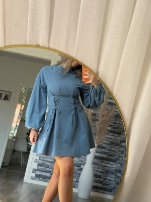 Blå kort klänning med snörning - Supersnygg blå klänning med ballongärmar och snörningsdetaljer i midjan. Klänningen har en kort, utsvängd kjol och hög hals. Perfekt för dig som vill ha en trendig och unik look.