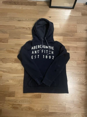 Mörkblå hoodie från Abercrombie & Fitch - Snygg mörkblå hoodie från Abercrombie & Fitch med vit text på bröstet och dragsko i huvan. Klassisk modell med känguruficka och mjukt material, perfekt för en avslappnad stil.