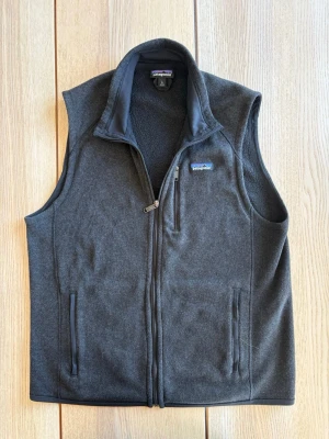 Patagonia fleeceväst - Snygg mörkgrå fleeceväst från Patagonia med hel dragkedja framtill och två fickor med dragkedja. Västen har hög krage och mjukt fleecefoder, perfekt för lager-på-lager. Klassisk Patagonia-logga på bröstet.