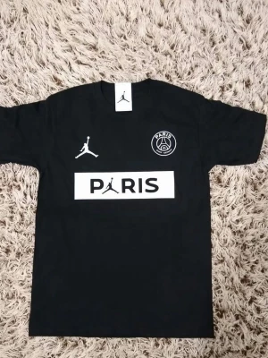Svart Paris Jordan T-shirt - Cool svart T-shirt från Jordan x Paris med vit logga och text på bröstet. Klassisk rund hals och korta ärmar. Perfekt för dig som gillar sportig streetstyle och vill sticka ut med en snygg och enkel design.