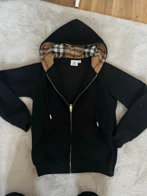 Svart hoodie med guld detaljer - En stilren svart hoodie med guldiga detaljer med snygga detaljer i luvan. Osäker på storlek men passa mig nästan perfekt som är xs. 
