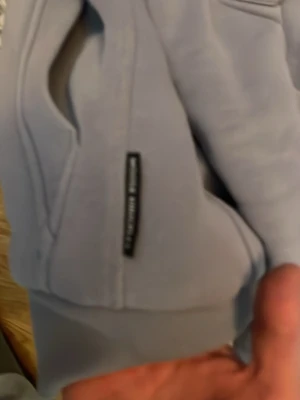 Ljusgrå hoodie från Moose Knuckles - Säljer en ljusgrå hoodie från Moose Knuckles med klassisk huva och magficka. Hoodien har ribbade muddar vid ärmslut och nederkant samt en liten svart Makia-logga på sidan av fickan. Perfekt för en chill och avslappnad stil.