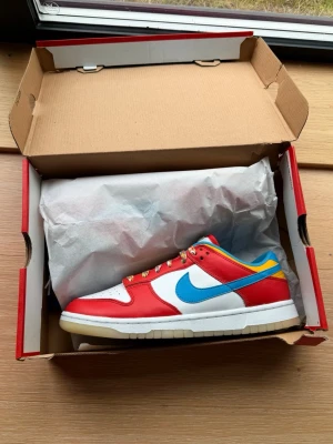 Nike Dunk Low Fruity Pebbles sneakers - Nike Dunk Low Fruity Pebbles sneakers med färgglada detaljer. Skorna har vit tåbox och sidor, röda overlays, blå swoosh och häl, samt gula och blå inslag runt kragen. Flerfärgade skosnören och Fruity Pebbles-tagg på plösen. Tillverkade i läder och textil.