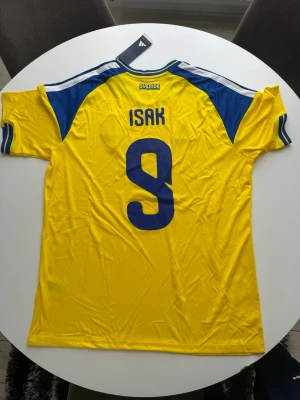 Sverige matchtröja Isak #9 Adidas - Gul och blå Sverige matchtröja från Adidas med Isak och nummer 9 på ryggen. Tröjan har blå detaljer på axlar och ärmar, samt det svenska landslagets emblem på bröstet. Tillverkad i lätt och ventilerande material, perfekt för fotboll eller träning.