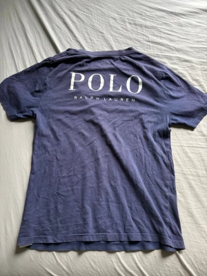 Blå t-shirt från Polo Ralph Lauren - Snygg blå t-shirt från Polo Ralph Lauren med stort vitt POLO-tryck på ryggen och klassisk logga på bröstet. T-shirten har rund hals och korta ärmar, perfekt för en avslappnad och stilren look.