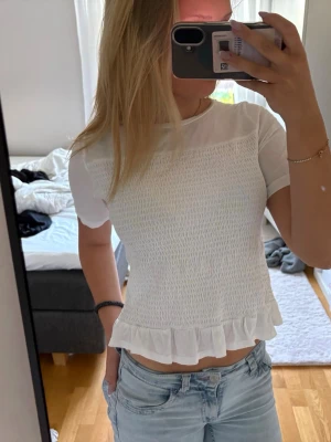 Vit topp med volangkant - Fin och enkel topp med korta ärmar och smockad kropp. Perfekt till nu inför sommaren. Den är använd men är i bra skick ändå. Priset kan diskuteras 