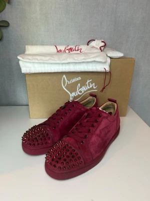 Sneakers Christian Louboutin - Snygga röda sneakers från Christian Louboutin i mocka med matchande röda nitar på tån och sidorna. Klassisk låg modell med rund tå och röda skosnören. Perfekta för dig som vill sticka ut med exklusiv design och edgy detaljer. 40 passar 41