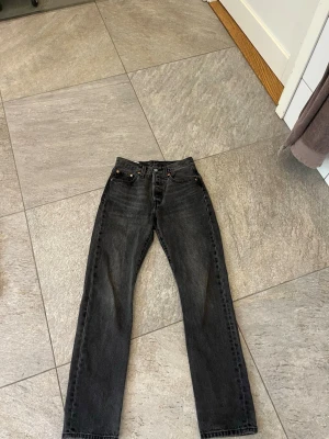 Levis 501 - Säljer ett par svarta raka jeans från Levi's med klassisk femficksdesign och knappgylf. Jeansen har en vintagekänsla och är tillverkade i slitstark denim. Perfekta för dig som gillar en tidlös och avslappnad stil.
