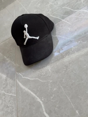 Svart keps med vit Jumpman-logga - Säljer en svart keps från Jordan med den klassiska vita Jumpman-loggan broderad framtill. Kepsen har böjd skärm och justerbart spänne bak. Tillverkad i bomullstyg för en bekväm och sportig look.