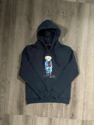 Mörkblå hoodie Polo Bear Ralph Lauren - Mörkblå hoodie från Ralph Lauren med den ikoniska Polo Bear-trycket på bröstet. Hoodien har huva med dragsko och ribbade muddar vid ärmar och nederkant. Tillverkad i mjukt bomullsmaterial för en skön och avslappnad look.