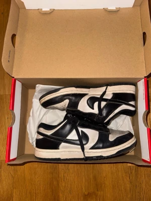Nike Dunk Low svart/vit sneaker - Säljer ett par klassiska Nike Dunk Low sneakers i svart och vitt läder i stl 39. Skorna har svart swoosh, svarta snören och vit tåbox med perforeringar. Tidlös modell som passar till det mesta och har en låg siluett. Kommer i originalkartong.