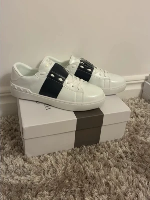 Valentino vita sneakers med svart band - Snygga vita sneakers från Valentino med ett brett svart band över vristen och guldfärgad logga på sidan. Skorna har vita snören, platt sula och är tillverkade i slätt läder. Perfekta för dig som vill ha en clean och exklusiv look.