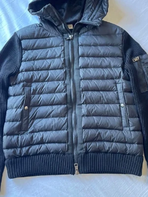Moncler Cardigan - Snygg svart pufferjacka med stickade ärmar och rygg samt vadderad framsida och huva. Jackan har dragkedja framtill, två fickor med tryckknappar och en extra ficka med dragkedja på. Riktigt bekväm och i gott skick. Skulle säga 7,5-8/10 då det finns fåtal noppriga prickar på vissa ställen och minimala skillnader, men inget man ser utan att leta efter det. Hör av vid minsta fundering.