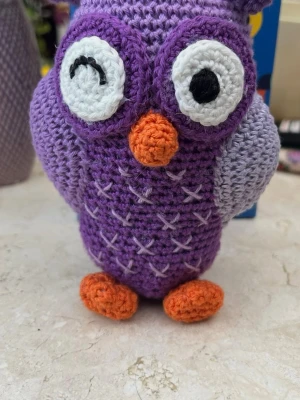 Virkad lila uggla prydnad - Söt handvirkad uggla i lila och ljuslila garn med orange näbb och fötter. Ugglan har stora vita ögon, varav ett blinkar, och broderade detaljer på magen. Perfekt som dekoration eller present.