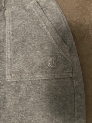 Grå mjukisbyxor från juicy - Mysiga grå mjukisbyxor från juicy med raka ben och bred resår i midjan. Byxorna har två stora fickor fram och en diskret broderad logga på ena fickan. Tillverkade i mjukt och stretchigt material som passar perfekt för chill dagar.