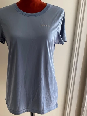 Ljusblå sportig t-shirt från Sport1 - Denna kortärmade träningströja i en lugn blå nyans är designad för att ge komfort under aktivitet. Tröjan har en rund halsringning och en diskret logotyp framtill. En reflekterande detalj löper längs ryggen, vilket kan öka synligheten. Passformen är anpassad för rörelsefrihet. Längd: 64 cm Bystvidd: 45 cm  Inga bilder på mig! Katter finns i hemmet!