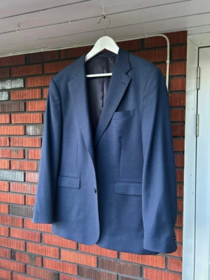 Klassisk blå kavaj med smal passform - Snygg blå kavaj med smal passform och klassiska slag. Kavajen har två framfickor med lock och en bröstficka. Perfekt för dig som vill ha en stilren och tidlös look. Materialet ser ut att vara tunn ull eller ullblandning. Endast använd 1 gång därmed i princip helt ny. Säljes under halva nypriset. 