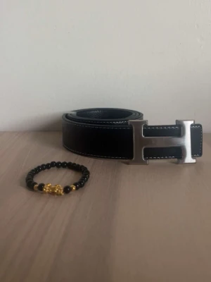 Svart skärp med H-spänne - Snyggt svart skärp med ett stort, silverfärgat H-spänne. Bältet har vita sömmar längs kanterna och är gjort i slätt skinn. Perfekt accessoar för att lyfta vilken outfit som helst.