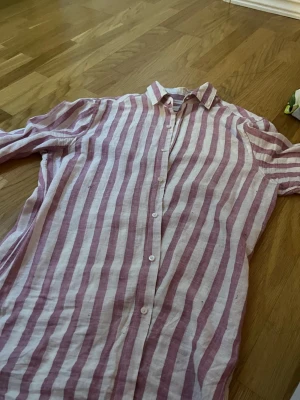 Randig linneskjorta från Massimo Dutti - Säljer en långärmad skjorta från Massimo Dutti i vitt och lila med breda ränder. Skjortan har klassisk krage, knäppning framtill och är tillverkad i svalt linne – perfekt för varma dagar. Snygg att styla både öppen och stängd.